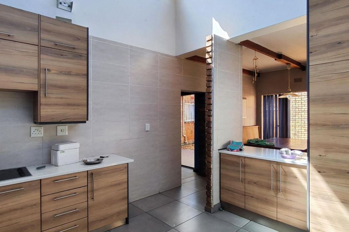 Modern kitchen featuring wood-look cabinetry, light countertops, ceramic tiling, and an exposed brick architectural detail, connecting to an adjacent living area.