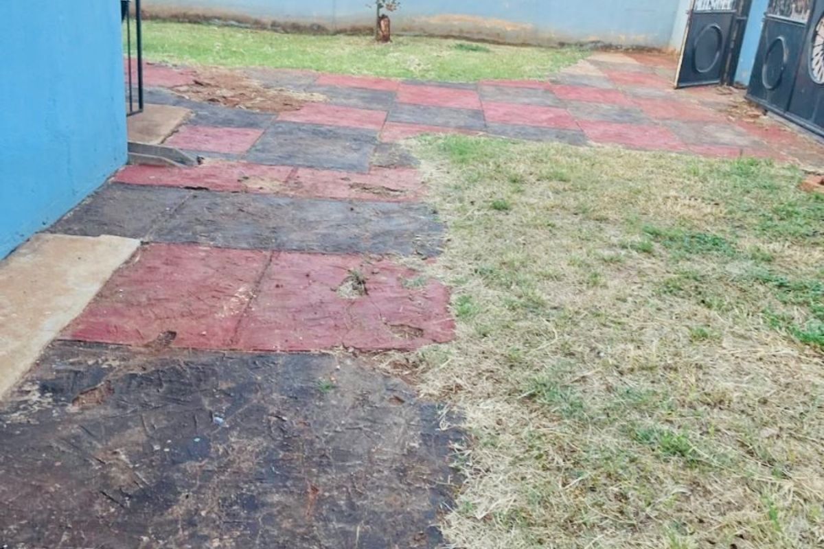 A rear garden area featuring a perimeter wall, metal gate, and a paved patio section in need of maintenance and landscaping.