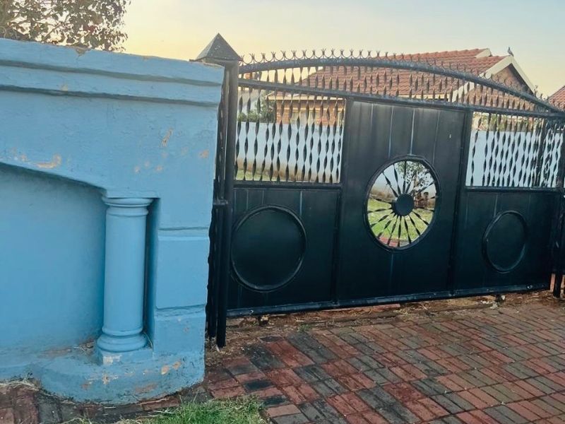 Front entrance featuring a decorative black metal driveway gate, masonry boundary wall with pillar detail, and brick paved driveway.