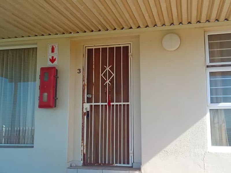 Exterior view of an apartment entrance featuring a security gate, fire safety equipment, and aluminium window frames under a corrugated roof overhang.