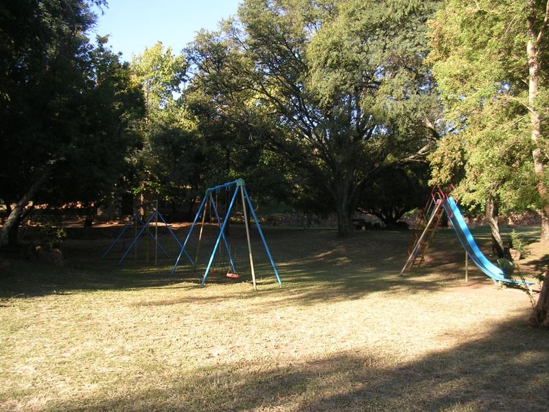 A spacious, tree-lined garden area featuring established playground equipment, including swings and a slide, set within a natural, grassy landscape.