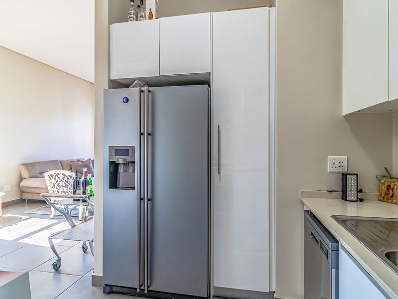 Modern kitchen area featuring sleek white cabinetry, stone countertops, a stainless steel sink, and integrated dishwasher, flowing into an open-plan living space.