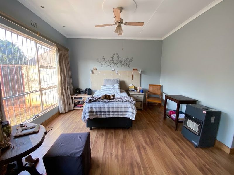 A spacious bedroom featuring laminate flooring, a ceiling fan, and large windows fitted with security bars, offering a functional and well-lit living space.