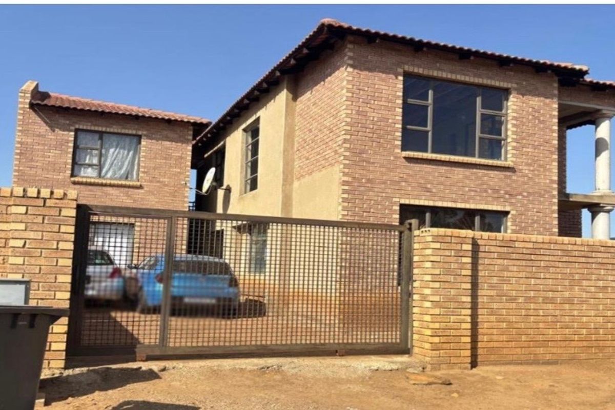 A two-story face-brick residential property featuring a secure gated entrance, paved driveway, and a covered balcony area.