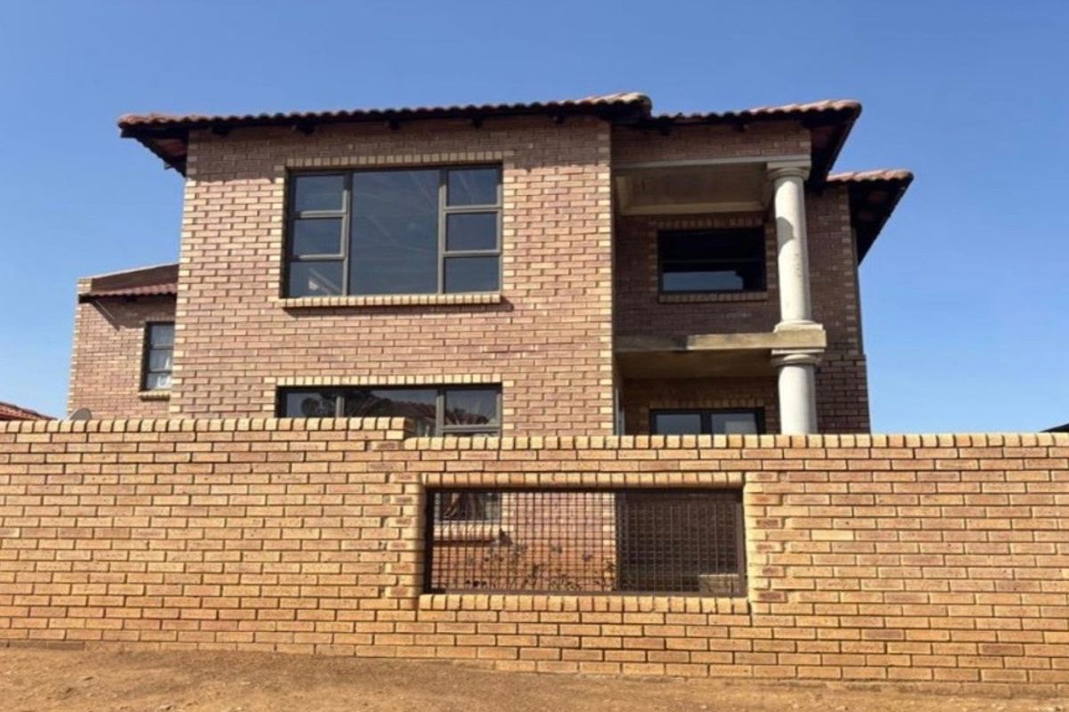 A double-storey face brick house featuring a tiled roof, aluminium window frames, and a secure perimeter wall with a metal gate.