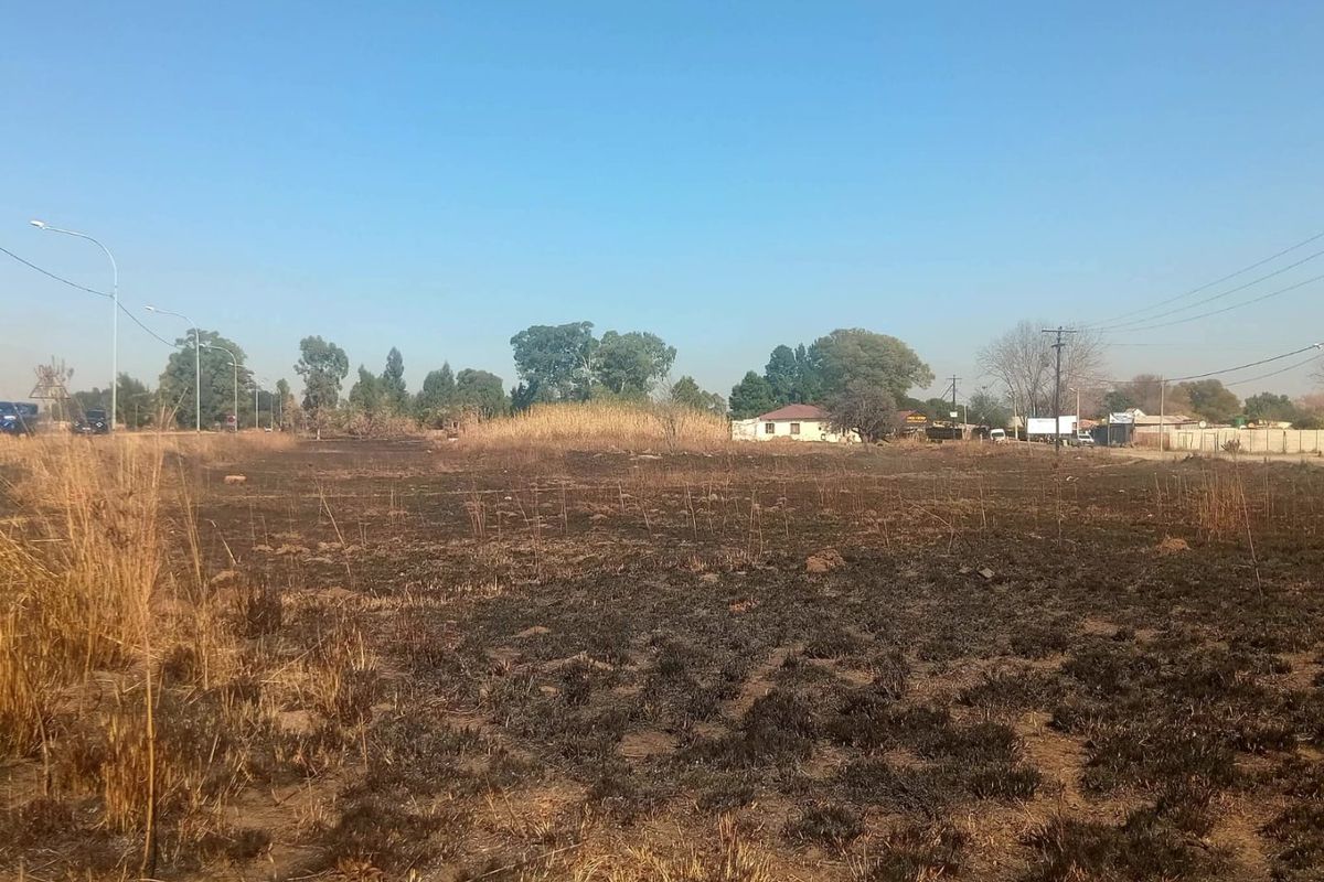 An open, undeveloped plot of land showing signs of recent fire damage to the vegetation, with distant residential structures and utility infrastructure visible.