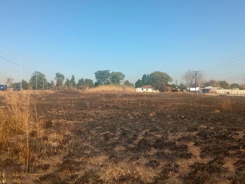 An open, undeveloped plot of land showing signs of recent fire damage to the vegetation, with distant residential structures and utility infrastructure visible.