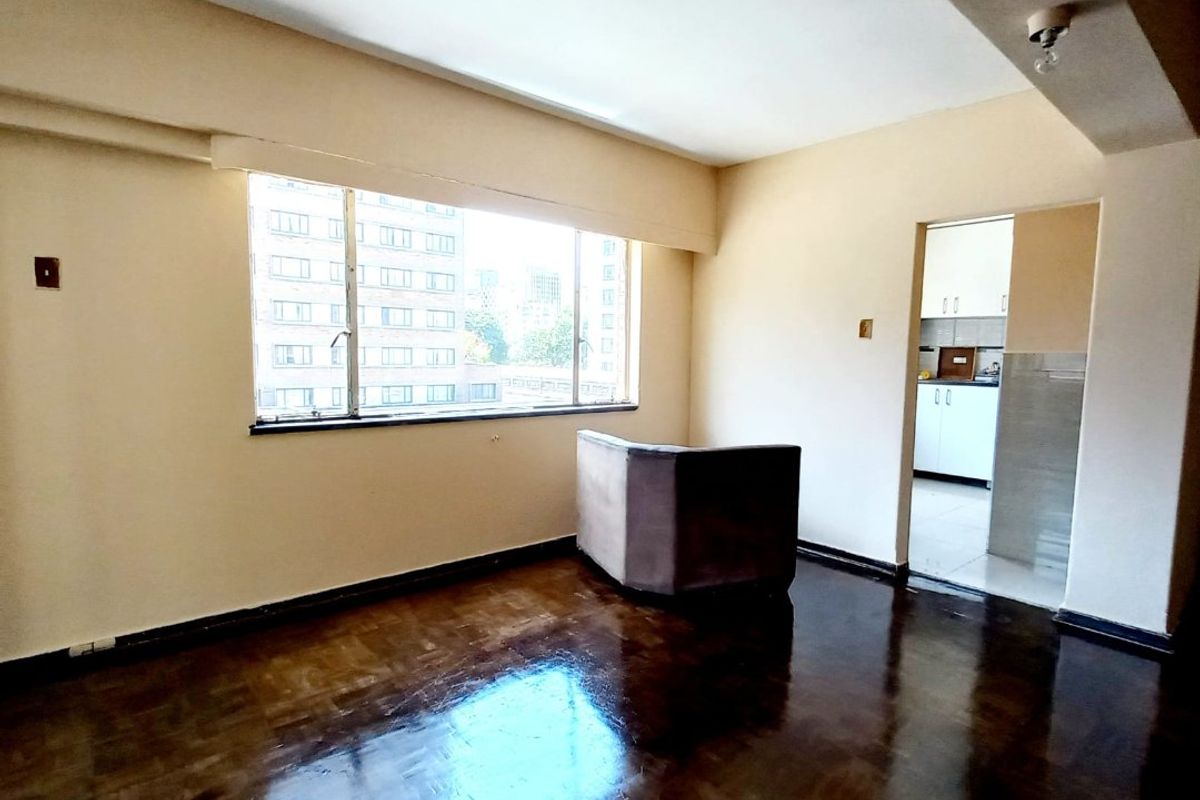 A spacious, empty living room featuring dark parquet flooring, a large window with natural light, and a view into the adjacent kitchen area.
