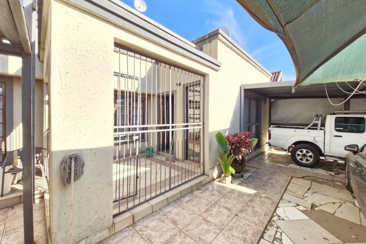 Rear exterior view featuring a tiled patio area, security gates, and paved driveway space with overhead shade sail protection.