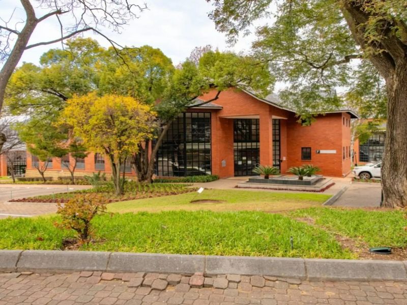 A professional exterior view of a commercial brick building featuring large glass windows, surrounded by a well-maintained garden and mature trees.