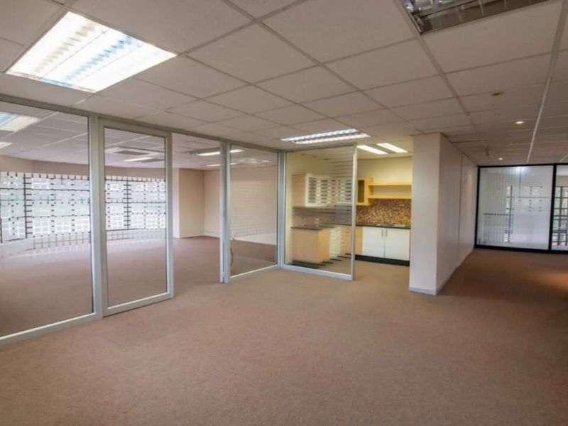 Open-plan commercial office space featuring glass partitions, suspended ceilings with recessed lighting, carpeted flooring, and a small integrated kitchenette area.