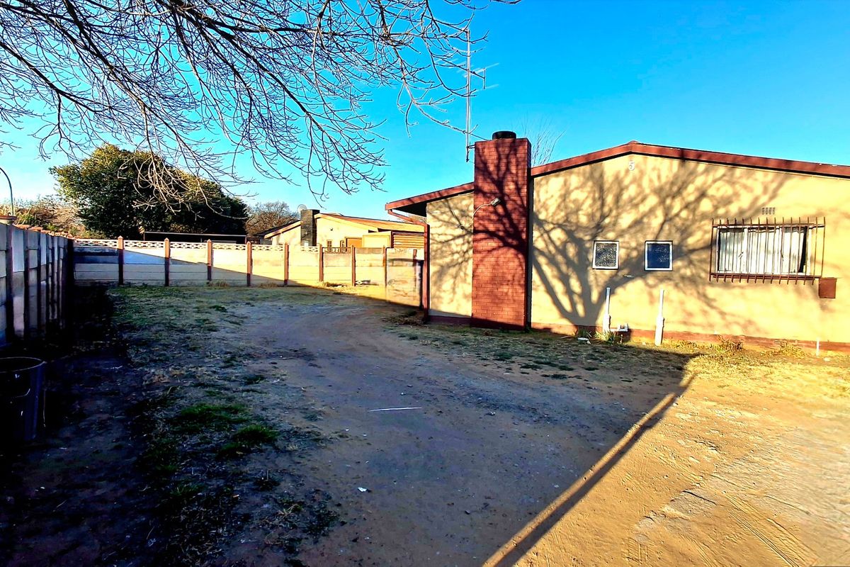 Rear exterior view of the property featuring a brick chimney, security bars on windows, and a large unpaved yard enclosed by a precast boundary wall.