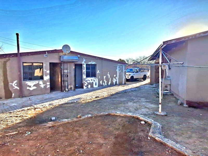 Rear exterior view of the property showing the building facade with visible plaster and paint deterioration, paved areas, and security features.