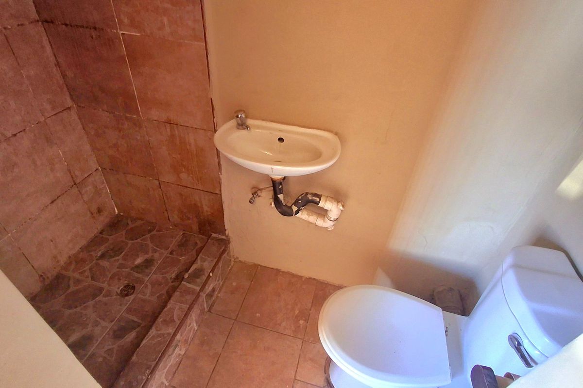 A compact bathroom featuring a shower enclosure, wall-mounted basin with exposed plumbing, and a standard toilet, requiring cosmetic updates and finish improvements.