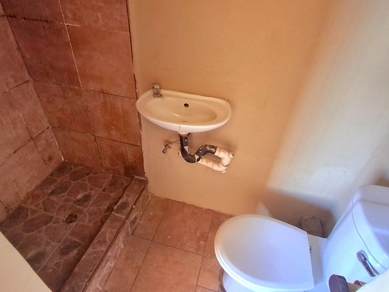 A compact bathroom featuring a shower enclosure, wall-mounted basin with exposed plumbing, and a standard toilet, requiring cosmetic updates and finish improvements.