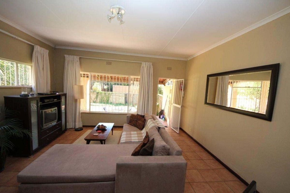 A spacious living room featuring tiled flooring, large windows providing natural light, and direct access to an outdoor area through a security gate.