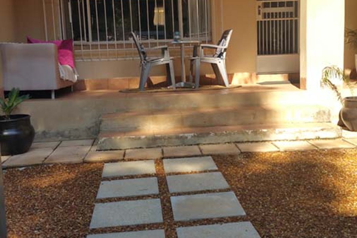 A covered rear patio area featuring tiled flooring, security bars on windows, and a paved walkway leading through the garden.