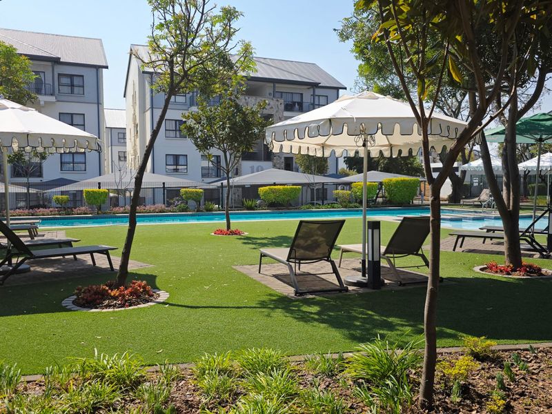 A well-maintained communal swimming pool area featuring artificial turf, paved lounging sections, and established landscaping within an apartment complex.