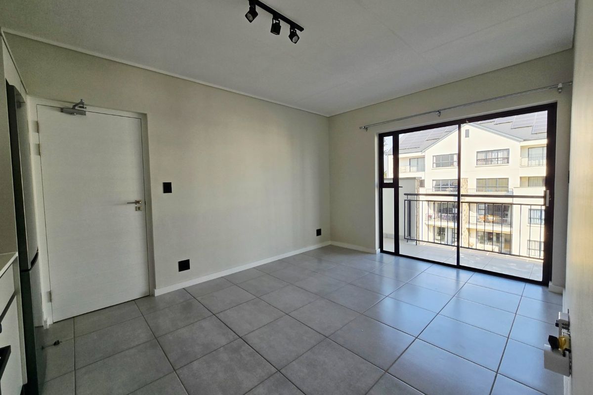 A bright, empty living area featuring neutral tiled flooring, modern track lighting, and a large sliding glass door providing access to a private balcony.