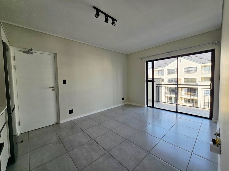 A bright, empty living area featuring neutral tiled flooring, modern track lighting, and a large sliding glass door providing access to a private balcony.
