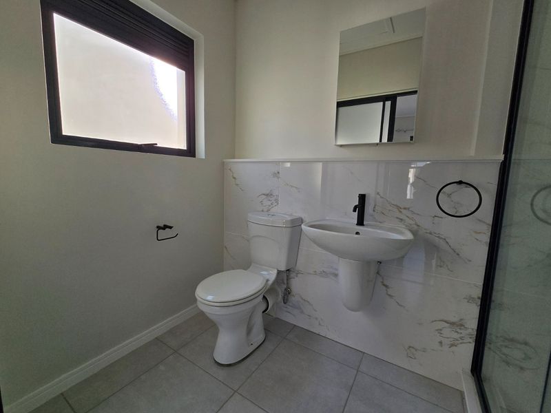 A modern, clean bathroom featuring a pedestal washbasin, toilet, marble-look wall tiling, and a shower enclosure with contemporary black fixtures.