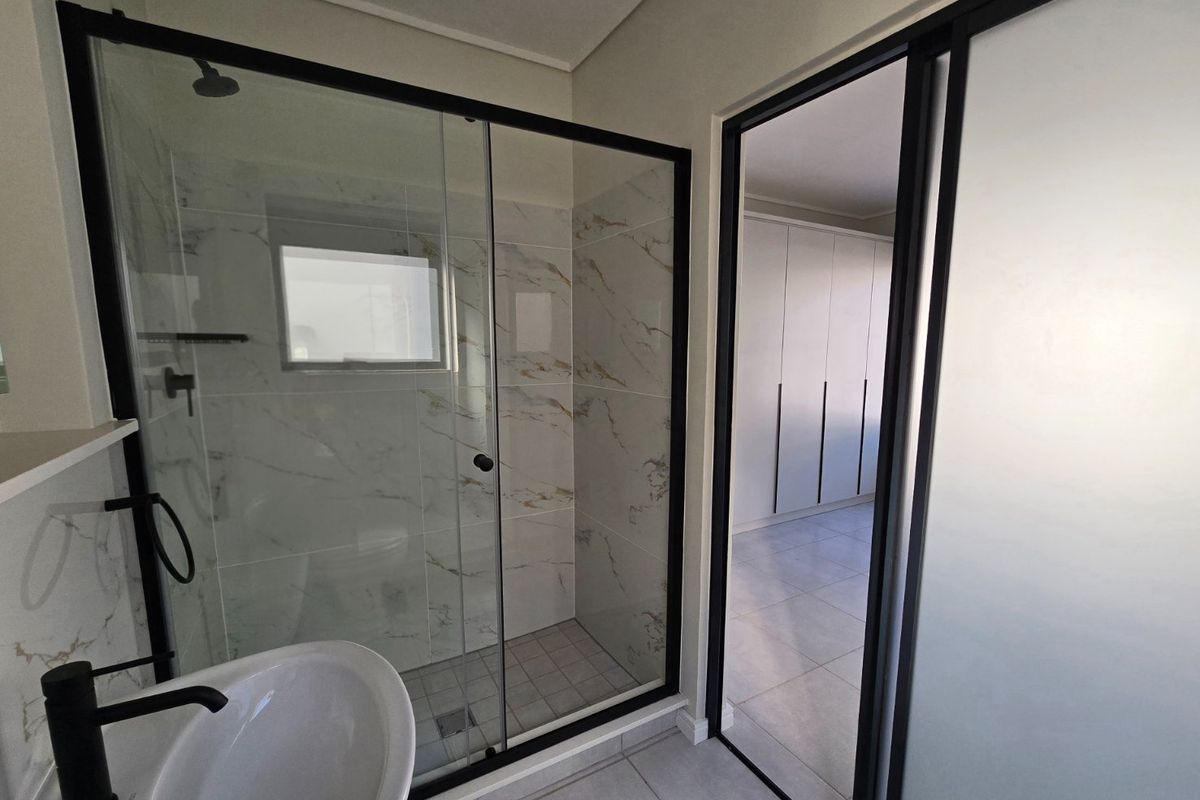 Modern bathroom featuring a walk-in shower with black-framed glass, marble-look tiling, and a sleek pedestal basin, adjacent to a room with built-in cupboards.