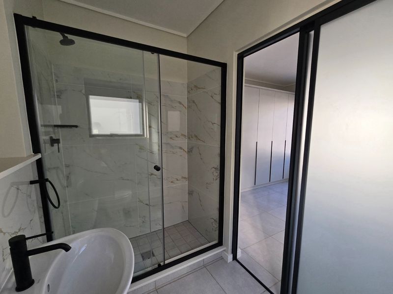 Modern bathroom featuring a walk-in shower with black-framed glass, marble-look tiling, and a sleek pedestal basin, adjacent to a room with built-in cupboards.