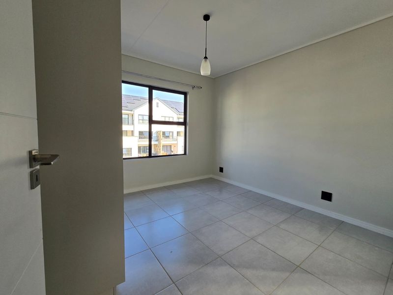 A bright, empty bedroom featuring neutral tiled flooring, a modern pendant light fixture, and a large window providing natural light.