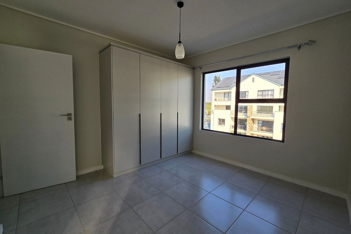A bright, empty bedroom featuring neutral tiled flooring, built-in cupboards, and a large window providing natural light.
