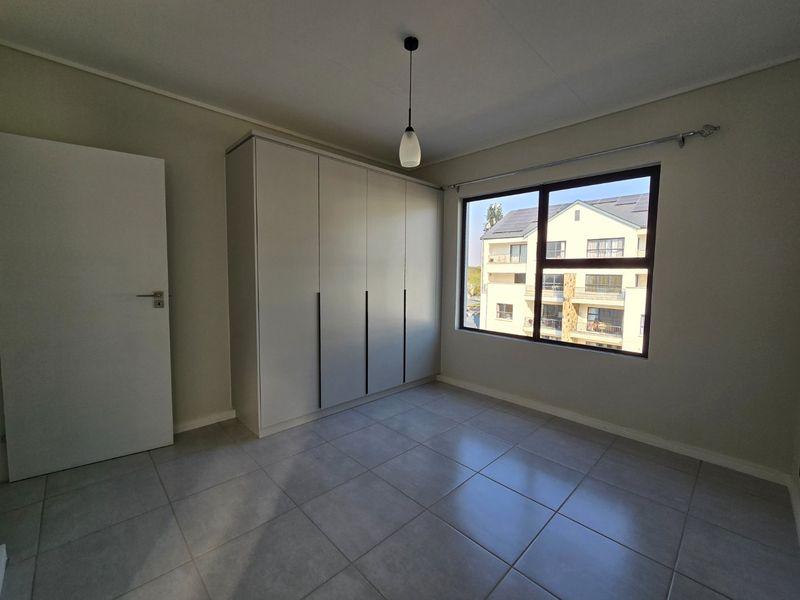 A bright, empty bedroom featuring neutral tiled flooring, built-in cupboards, and a large window providing natural light.