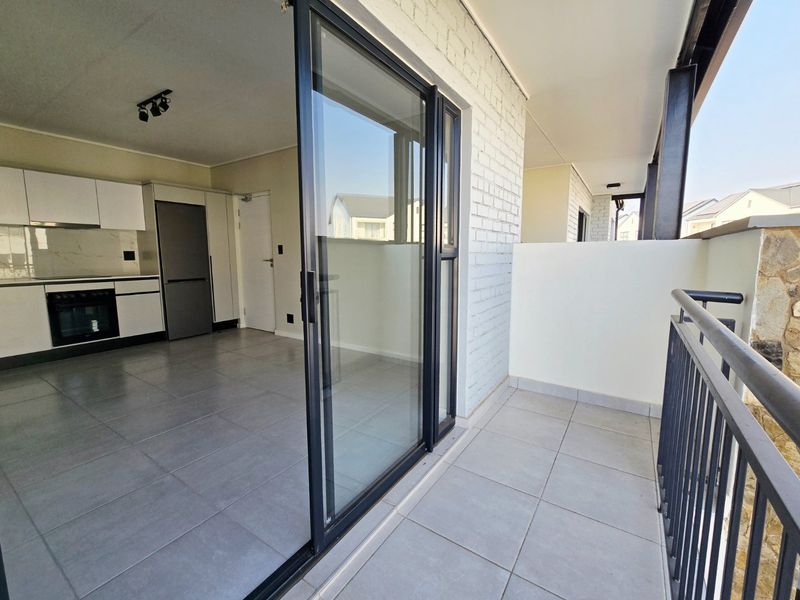 Modern open-plan apartment featuring a sleek kitchen with built-in appliances, flowing seamlessly onto a private balcony with metal balustrades and tiled flooring.