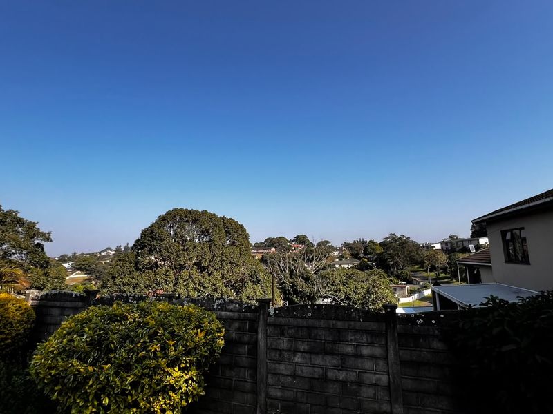 An elevated view overlooking the surrounding neighbourhood, featuring a concrete block boundary wall and mature trees under a clear blue sky.