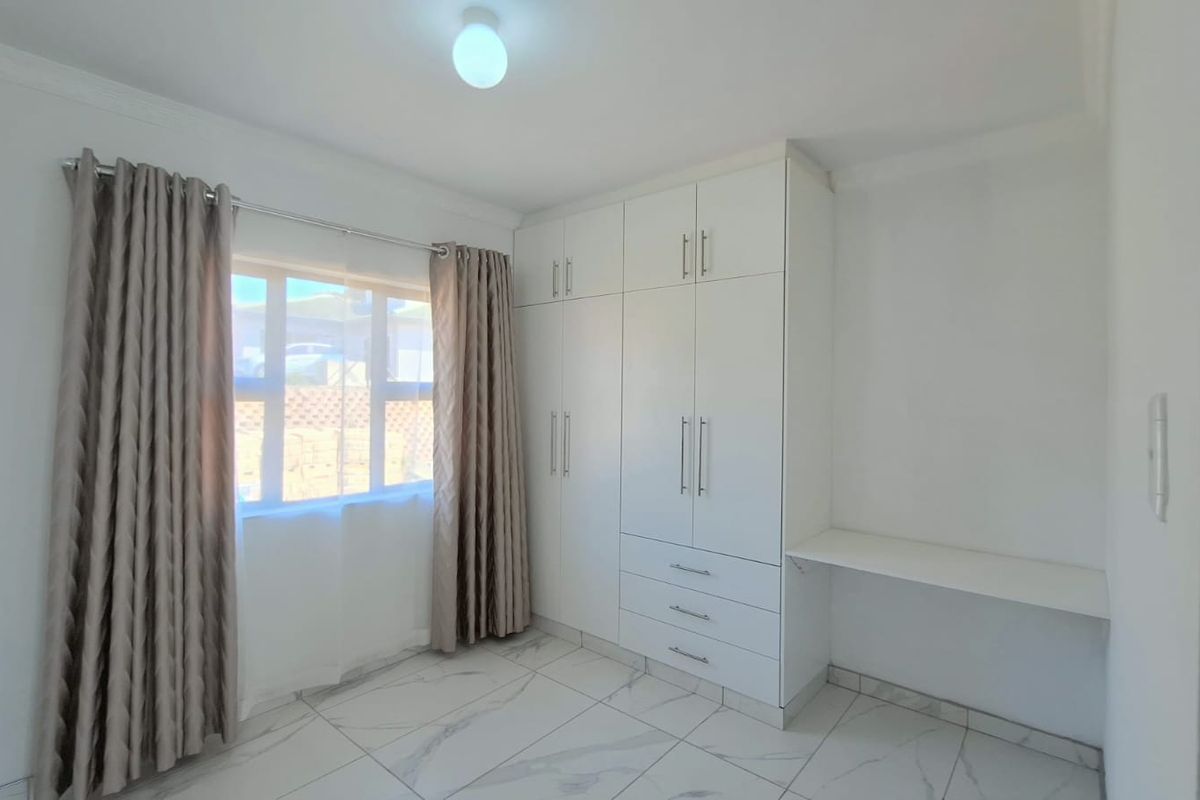 A bright bedroom featuring modern white built-in wardrobes, a built-in desk, and clean tiled flooring.