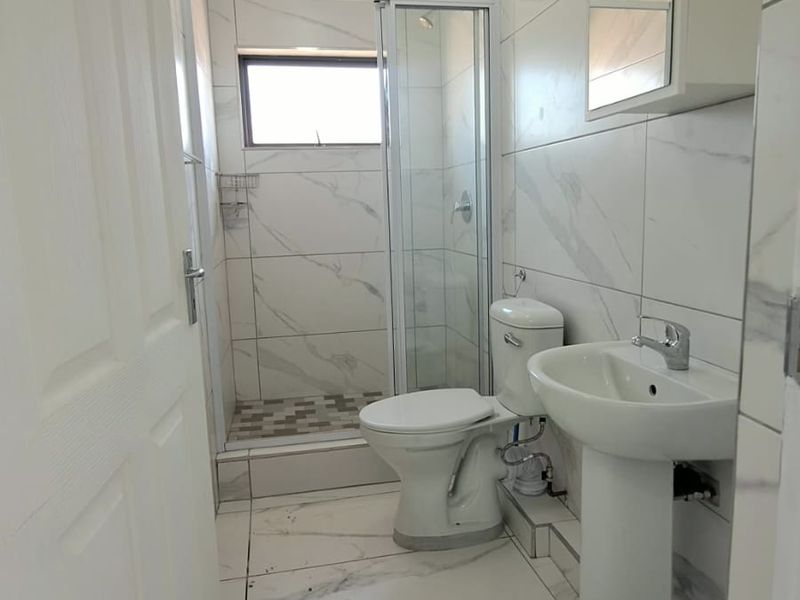 A compact bathroom featuring marble-look tiling, a pedestal basin, a toilet, and a glass-enclosed shower unit.