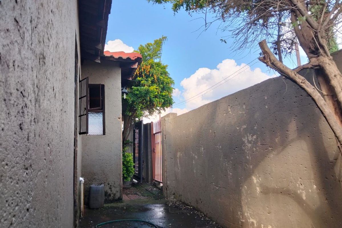 A narrow side passage of the property featuring a boundary wall, metal gate, and exterior wall access with visible weathering.