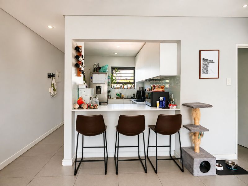 A modern kitchen featuring a breakfast bar, sleek white cabinetry, glass splashback, and recessed lighting, seamlessly integrated into the open-plan living area.