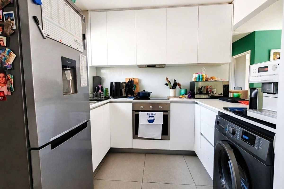 A compact, modern kitchen featuring white cabinetry, integrated appliances, a glass splashback, and neutral floor tiles.