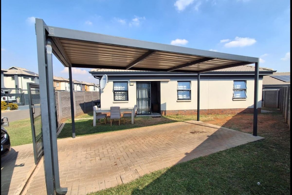 A rear exterior view featuring a paved driveway with a modern metal carport, brick boundary wall, and aluminium window frames.