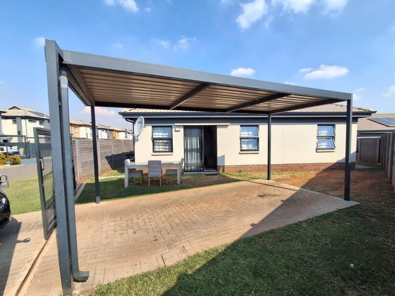 A rear exterior view featuring a paved driveway with a modern metal carport, brick boundary wall, and aluminium window frames.