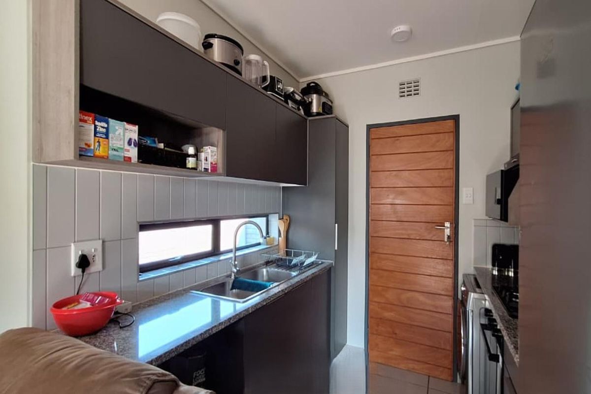 Compact kitchen featuring modern dark cabinetry, granite countertops, a stainless steel sink, and a tiled backsplash with a horizontal window for natural light.