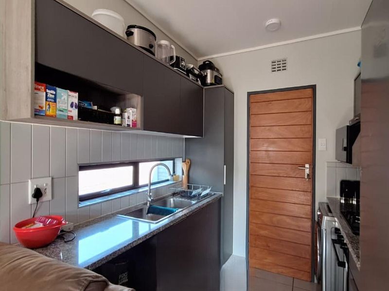 Compact kitchen featuring modern dark cabinetry, granite countertops, a stainless steel sink, and a tiled backsplash with a horizontal window for natural light.