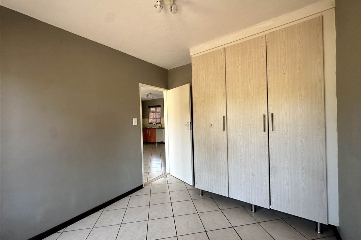 A clean, empty bedroom featuring light wood-finish built-in cupboards and neutral tiled flooring, with a view through to the kitchen area.
