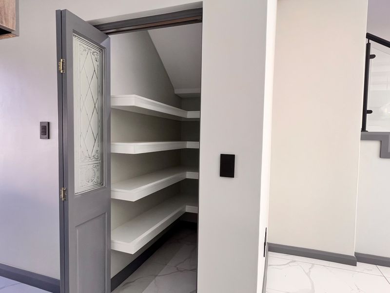A clean, modern walk-in pantry or scullery featuring built-in white shelving, a decorative glass door, and sleek marble-look floor tiles.
