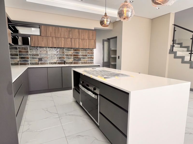 Modern kitchen featuring a central island with built-in oven, quartz countertops, decorative tiled backsplash, and an adjacent staircase with glass balustrade.