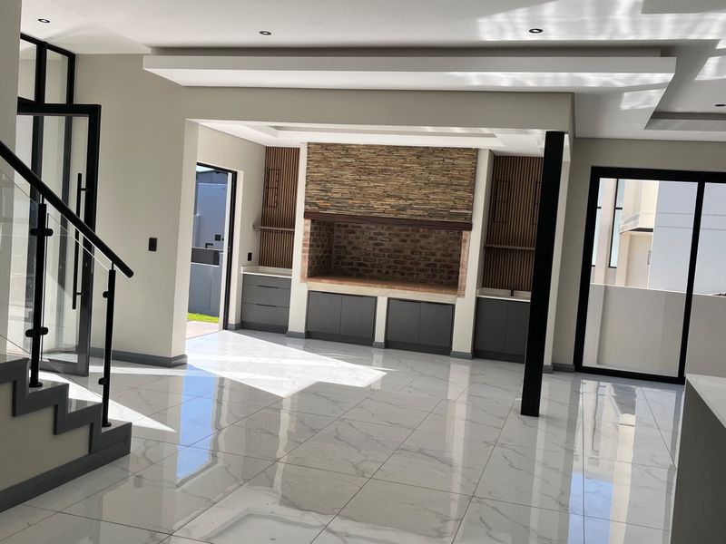 Spacious open-plan living area featuring a built-in braai with stone cladding, modern porcelain tiles, and a sleek glass staircase balustrade.