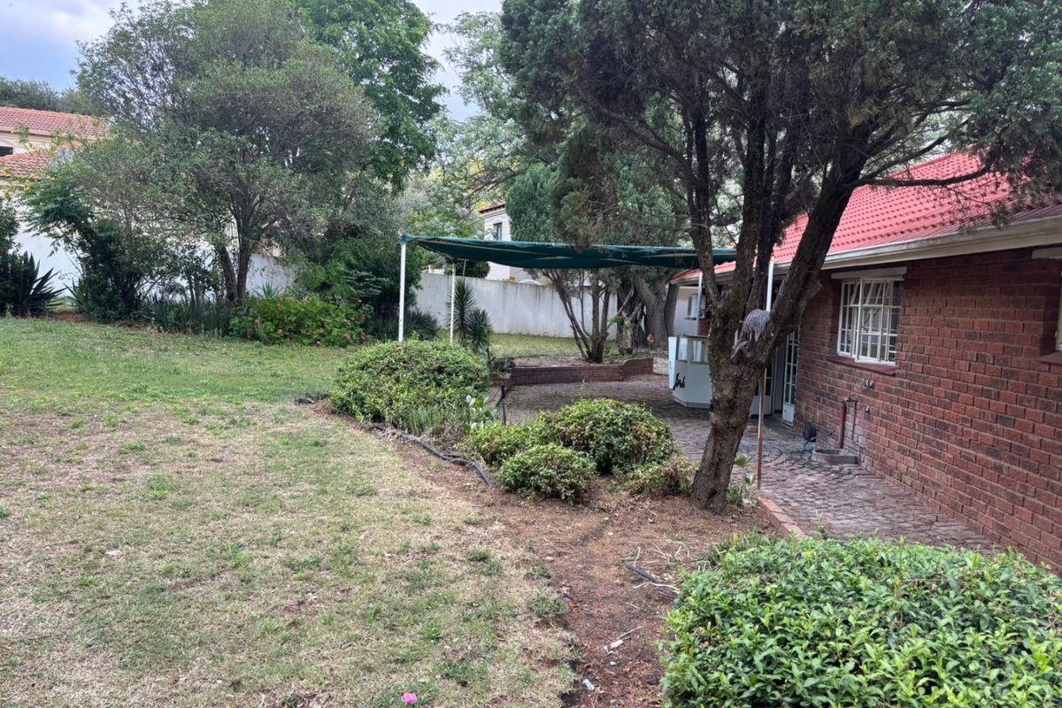 A rear view of the property featuring a brick facade, tiled roof, paved patio area with a shade sail, and a spacious garden.