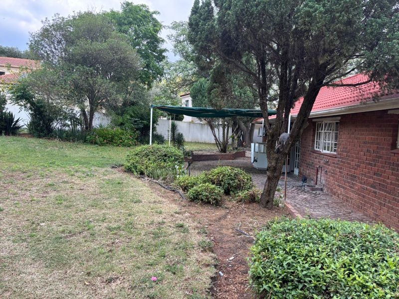 A rear view of the property featuring a brick facade, tiled roof, paved patio area with a shade sail, and a spacious garden.