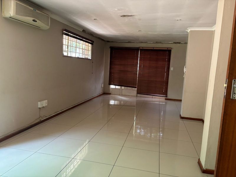An empty living room featuring tiled flooring, a wall-mounted air conditioning unit, and window blinds, showing signs of ceiling and wall finish wear.
