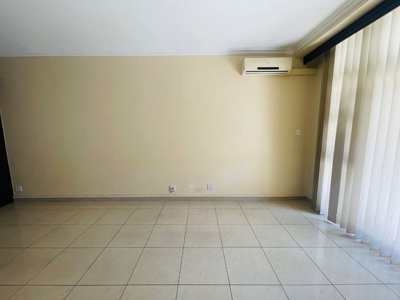 A bright, empty living room featuring neutral tiled flooring, vertical blinds, and a wall-mounted split-system air conditioning unit.