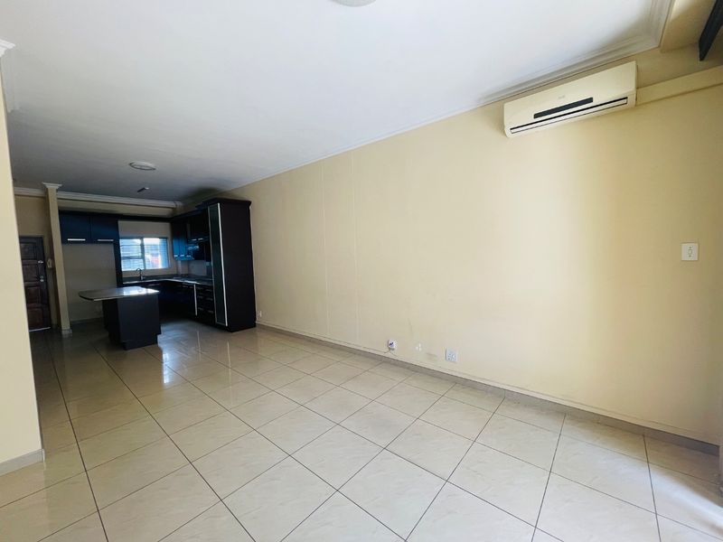 An open-plan living and kitchen area featuring tiled flooring, built-in dark cabinetry, a kitchen island, and a wall-mounted air conditioning unit.